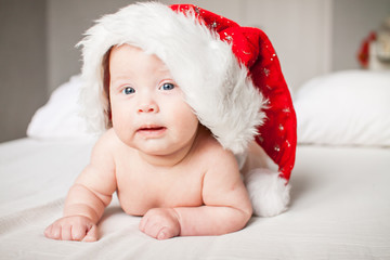 Cute funny infant baby in a big Santa Claus hat looking at camera on a white bed. New Year. Cheerful kid. Copyspace