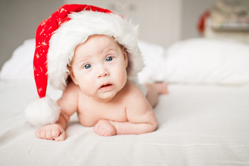 Cute funny infant baby in a big Santa Claus hat looking at camera on a white bed. New Year. Cheerful kid. Copyspace