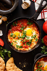 Shakshuka traditional Middle Eastern dish based on tomatoes, peppers and eggs in a cast iron skillet on a black background, top view