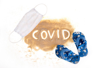 Sand on a white background. The inscription COVID on the hub, a protective mask around the sand and flip-flops. A vision of summer in 2020 during the Coronavirus pandemic. Summer in Covi-19