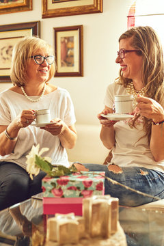 Beautiful adult daughter is giving flowers to her senior mom while drinking coffee at home.