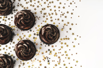 Chocolate cupcakes with chocolate icing and sprinkled gold sparkles on white background with golden star shaped confetti. Holiday concept. Mockup for your design. Flat lay style. Place for text.