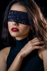 Portrait of beautiful young woman tying her eyes with black scarf on black background