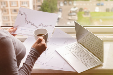 Woman freelancer working on laptop with graphics and charts printed on the paper standing near the window of her apartment. Girl drinking coffee early in the morning. remote work