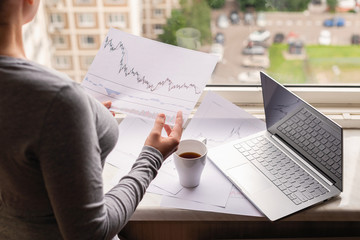 Woman freelancer working on laptop with graphics and charts printed on the paper standing near the window of her apartment. Girl drinking coffee early in the morning. remote work