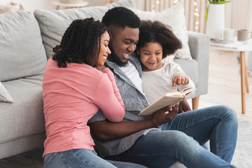 Happy black family reading book together, sitting on floor in living room