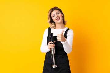 Girl using hand blender isolated on yellow background with thumbs up because something good has happened