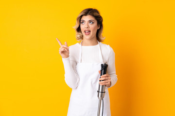 Girl using hand blender isolated on yellow background surprised and pointing finger to the side