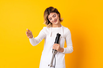 Girl using hand blender isolated on yellow background presenting and inviting to come with hand