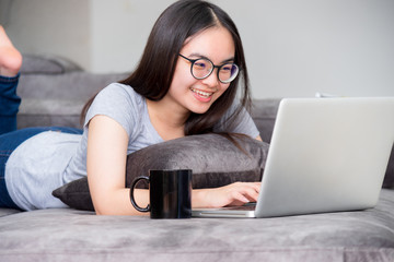 Portrait beautiful Asian young woman lying relax work with a laptop, Cute girl teens looking at the monitor smile happily on the sofa working remotely with an internet computer communication from home