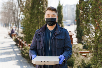 Fototapeta premium Young caucasian courier in black medical mask and blue gloves with one box of pizza in his hands. Quarantine for stopping pandemic of coronavirus Covid 19