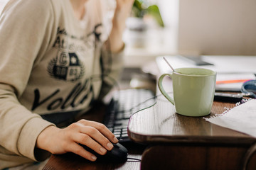 selective focus: young woman working in an online call center arranged in her room by the window. Home environment, she drinks coffee and simultaneously works on computer