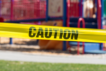 Close-up of yellow cordon tape with word caution, with public park in background