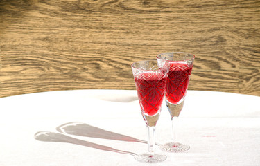 Two crystal glasses with red wine. Glass goblets with alcohol on background of wooden wall. White linen tablecloth. Direct sunlight, long shadows. Selective focus image. Copy space.