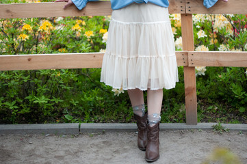 stylish girl in a light skirt and brown cowboy boots near a wooden fence with flowers in the garden