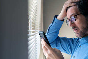 Man standing by window reacting to what he is viewing on his mobile phone.