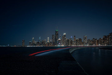 Chicago city skyline at night with a long red and white light trail left by an electric skateboard on a curved pier at North Avenue Beach creating a beautiful cityscape image or background.