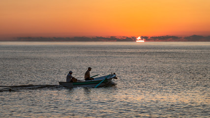 Naklejka premium Two fishermen passing by the boat with the sun set at sunset on the horizon. They're returning home to their prey.
