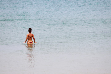 Sea vacations, slim girl in red thong bikini standing waist deep in blue water, rear view. Concept of swimming, sexy body, empty beach at quarantine during covid-19 coronavirus pandemic