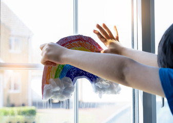 Kid putting painting colouful rainbow with cotton wool on white paper in the window during Covid-19 quarantine at home.Stay at home Social media campaign for coronavirus prevention concept