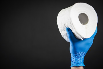 Close-up detail on a man's hand in a blue rubber glove as he holds a white roll of toilet paper on the black background. The topic is about Corona virus - COVID 19.