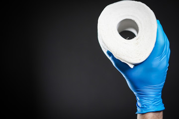 Close-up detail on a man's hand in a blue rubber glove as he holds a white roll of toilet paper on the black background. The topic is about Corona virus - COVID 19.