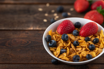 Cornflakes with blueberries and raspberries in white bowl on dark wooden desk.