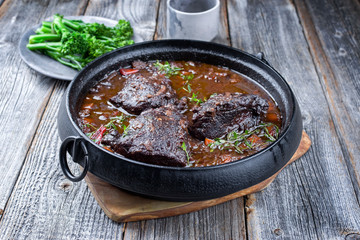 Traditionelle deutsche geschmorte Ochsenbäckchen  in brauner Rotwein Sauce mit Karotten und Broccoli angeboten als closeup in einem Modern Design Schmortopf auf einem alten rustikalen Board
