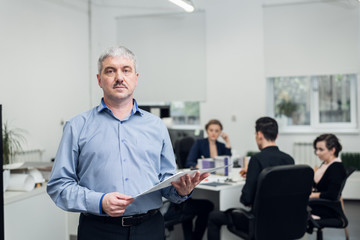 An aged office employee in a blue shirt holding some technical documents, his colleagues are having a meeting in the background