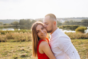 Beautiful young woman in red dress with her boyfriend outside on natural green background with sunlight. Handsome young man kiss her girlfriend .
