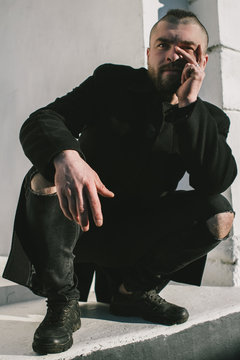 A bearded man in a black coat and ripped jeans squats, clasped his hand on his chin, stares intently at the camera. The guy in black is sitting against a white wall. Men's style and fashion.