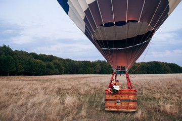 Couple in love staying in balloon basket with bouquet of white roses on a wide field. Handsome man confessed in love to his beautiful girlfriend in air balloon flight. Original proposal of marriage
