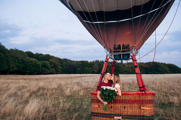 Couple in love kissing in ballon with bouquet of white roses on a wide field. Handsome man confessed in love to his beautiful girlfriend in air balloon flight . Original proposal of marriage