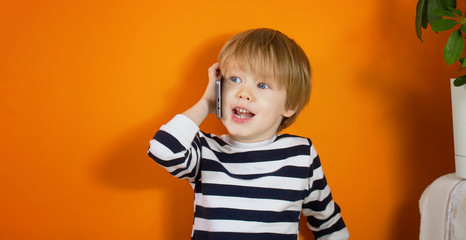Little boy in striped t-shirt is smiling, talking by smartphone, looking at side, posing against orange studio background. Happy childhood, gadget, advertising. Close up, copy space