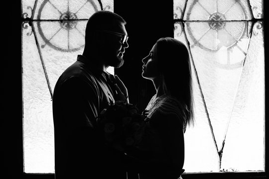 portrait of a romantic couple in a backlight from a window or door, silhouette of a couple in a doorway with a backlight, couple of lovers groom and bride at the window, girl holding flowers in hands - Powered by Adobe