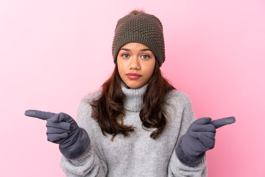 Young Colombian girl with winter hat over isolated pink wall pointing to the laterals having doubts