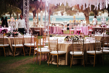 International Wedding outdoor celebration EVENING party under palm trees. Served tables on green area in hotel. Landyard. Beige and pink colors. Close-up and wide angle. - 341813465