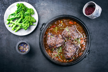 Traditional German braised beef cheeks in brown red wine sauce with carrots and broccoli offered as top view in a modern design stewpot on an old rustic board - obrazy, fototapety, plakaty