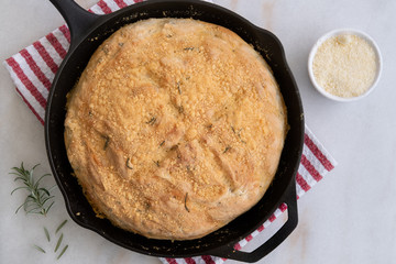 Just out of the oven, No Knead Rosemary Parmesan Skillet Bread, on a kitchen towel, with fresh rosemary and parmesan cheese