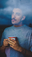 Boy holding cup of coffee, indoors, while looking out the window thinking looking outside.