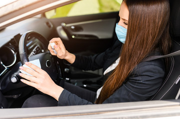 The girl in the car with a medical mask on a face is disinfecting the steering wheel with an...
