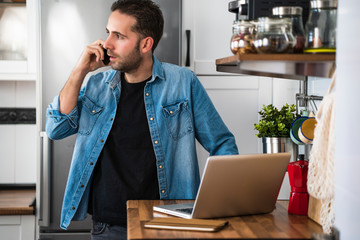 Man talking on his smartphone and using laptop in his home kitchen. Working at home and staying at home.