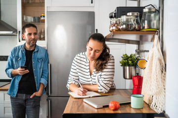 Young Man and Woman Couple talking and making the shopping list in their kitchen at home. Working at home and staying at home.