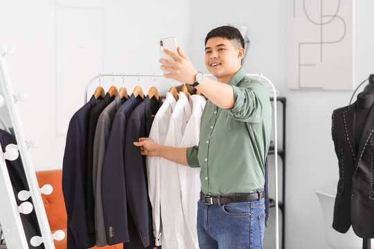 Male Asian stylist taking selfie near rack with clothes in studio