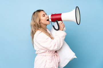 Teenager Russian girl in pajamas isolated on blue background shouting through a megaphone