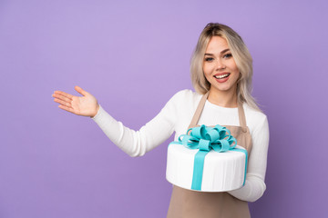 Young pastry chef holding a big cake over isolated purple background extending hands to the side for inviting to come