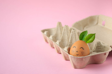a chicken egg with a cute painted face on it, lying in an egg box on a pink background, a festive...