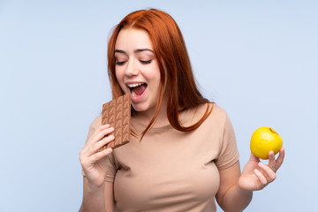 Redhead teenager girl over isolated blue background taking a chocolate tablet in one hand and an...
