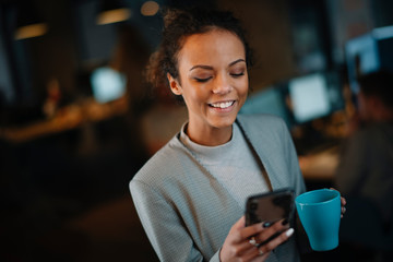 Young businesswoman using phone and drinking coffee in office.	