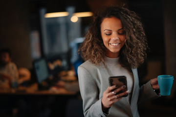 Young businesswoman using phone and drinking coffee in office.	